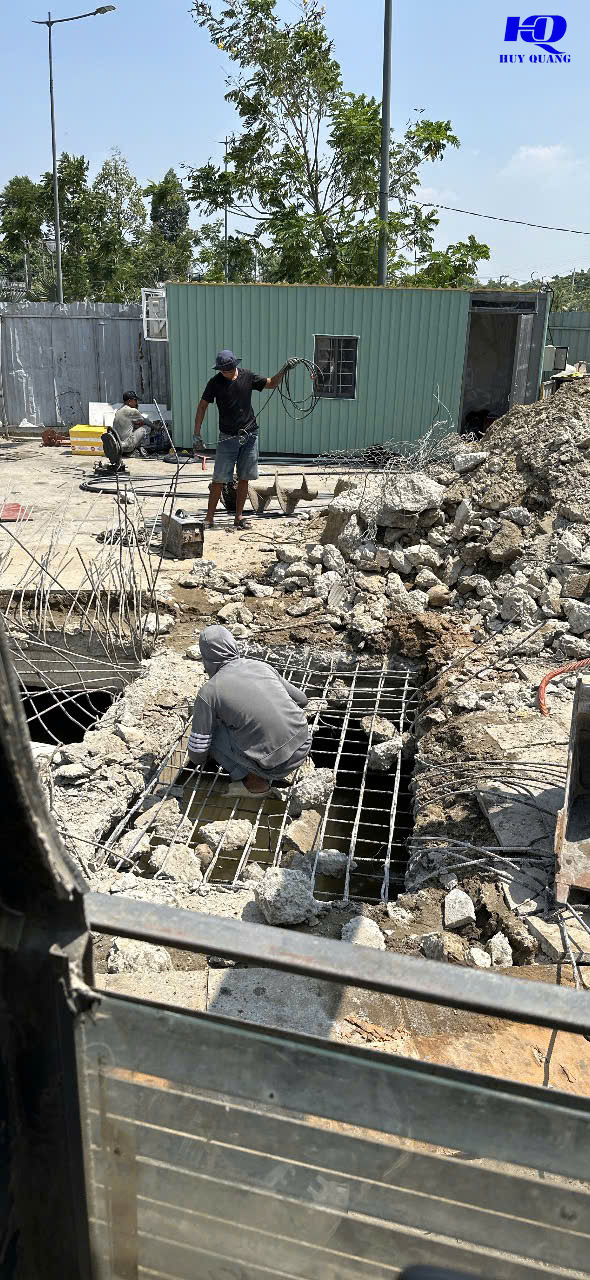 Công nhân thu gom dây cáp và cốt thép từ hố móng bê tông vừa đục phá, mảnh bê tông vỡ chất thành đống xung quanh, một công nhân khác cúi xử lý lưới thép dưới hố