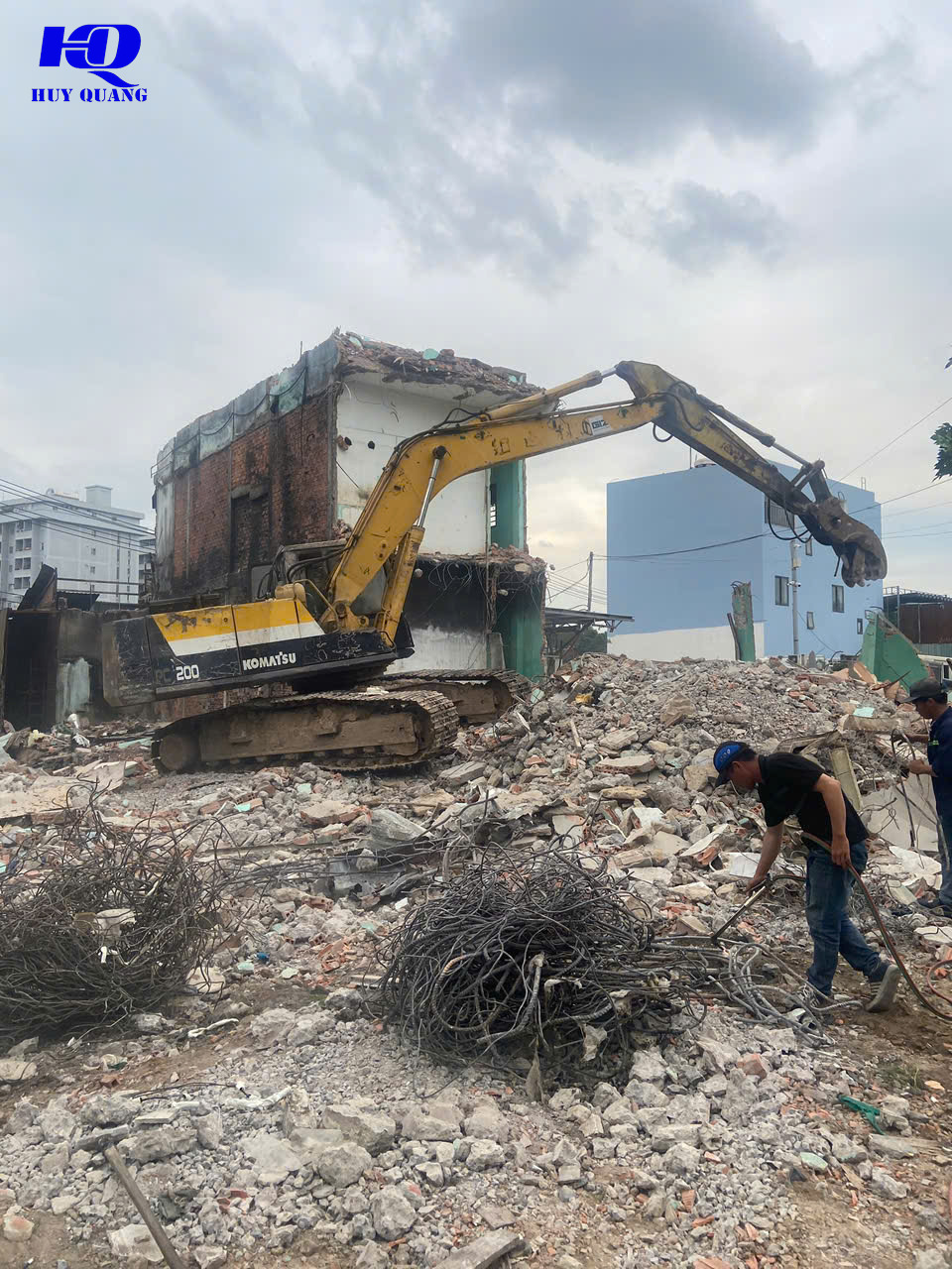 Máy đào Komatsu phá dỡ nhà phố lộ khung gạch đỏ bên trong, xà bần và cuộn dây điện thu hồi nằm phía trước, hai công nhân đang xử lý bên cạnh