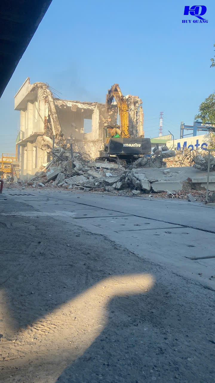 Máy đào Komatsu phá dỡ công trình bê tông nhiều tầng, phần kết cấu còn lại lộ khung thép và mảng tường trắng, khói bụi bốc lên từ khu vực đang thi công