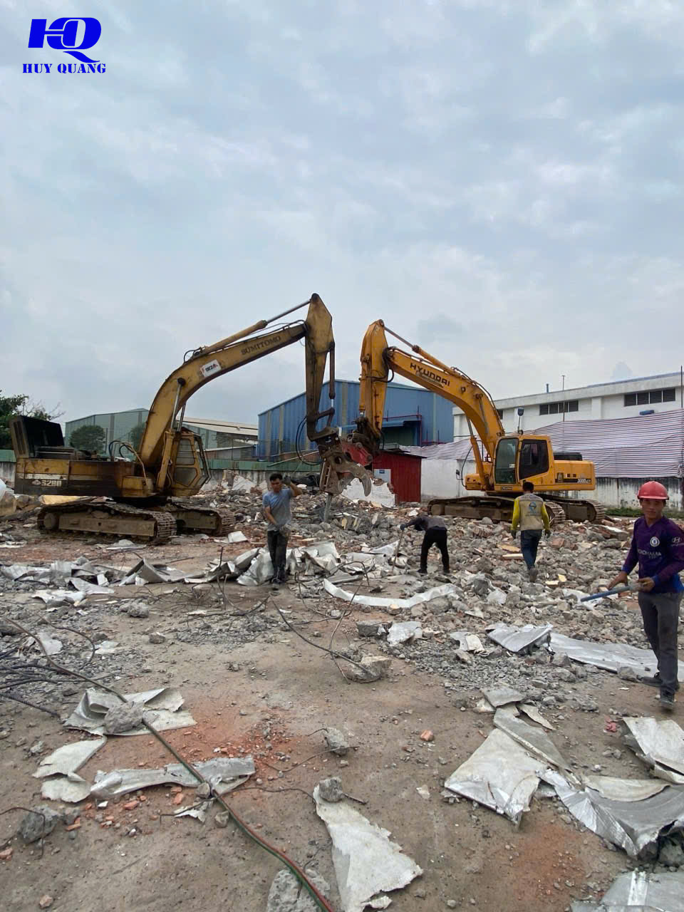 Hai máy đào phối hợp phá dỡ khung nhà xưởng, một máy gắn đầu kẹp thủy lực giật đổ mảng tường, công nhân đứng gần đống xà bần và tôn vụn phía trước