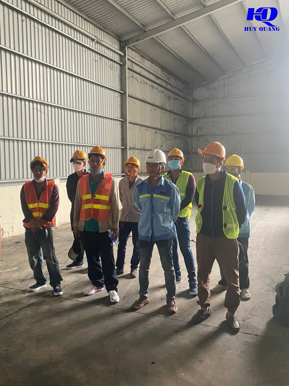 Nhóm công nhân và kỹ thuật viên đội mũ bảo hộ đứng họp trong nhà xưởng khung thép còn nguyên vẹn trước khi khảo sát vật tư thu mua