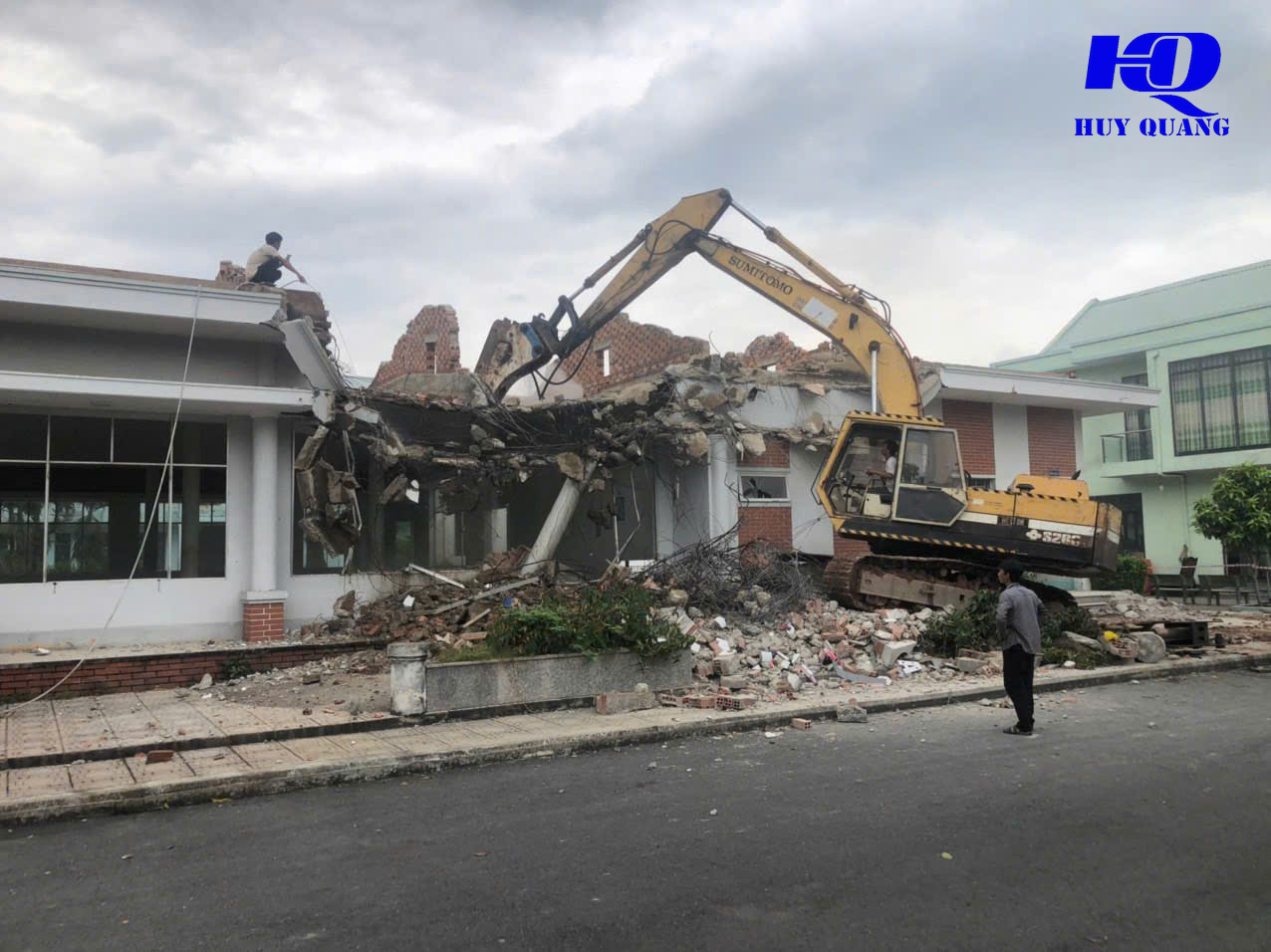 Máy đào phá dỡ phần mái và tường gạch của một căn biệt thự có sân vườn phía trước, xà bần đổ xuống lối đi lát gạch cạnh bồn cây