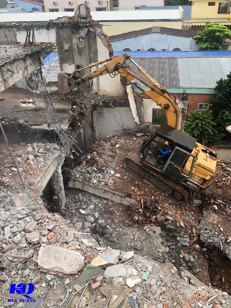 Máy đào xử lý phần kết cấu bê tông của nhà phố nhiều tầng nhìn từ trên cao, cốt thép lộ ra tại các mảng tường và sàn còn sót lại