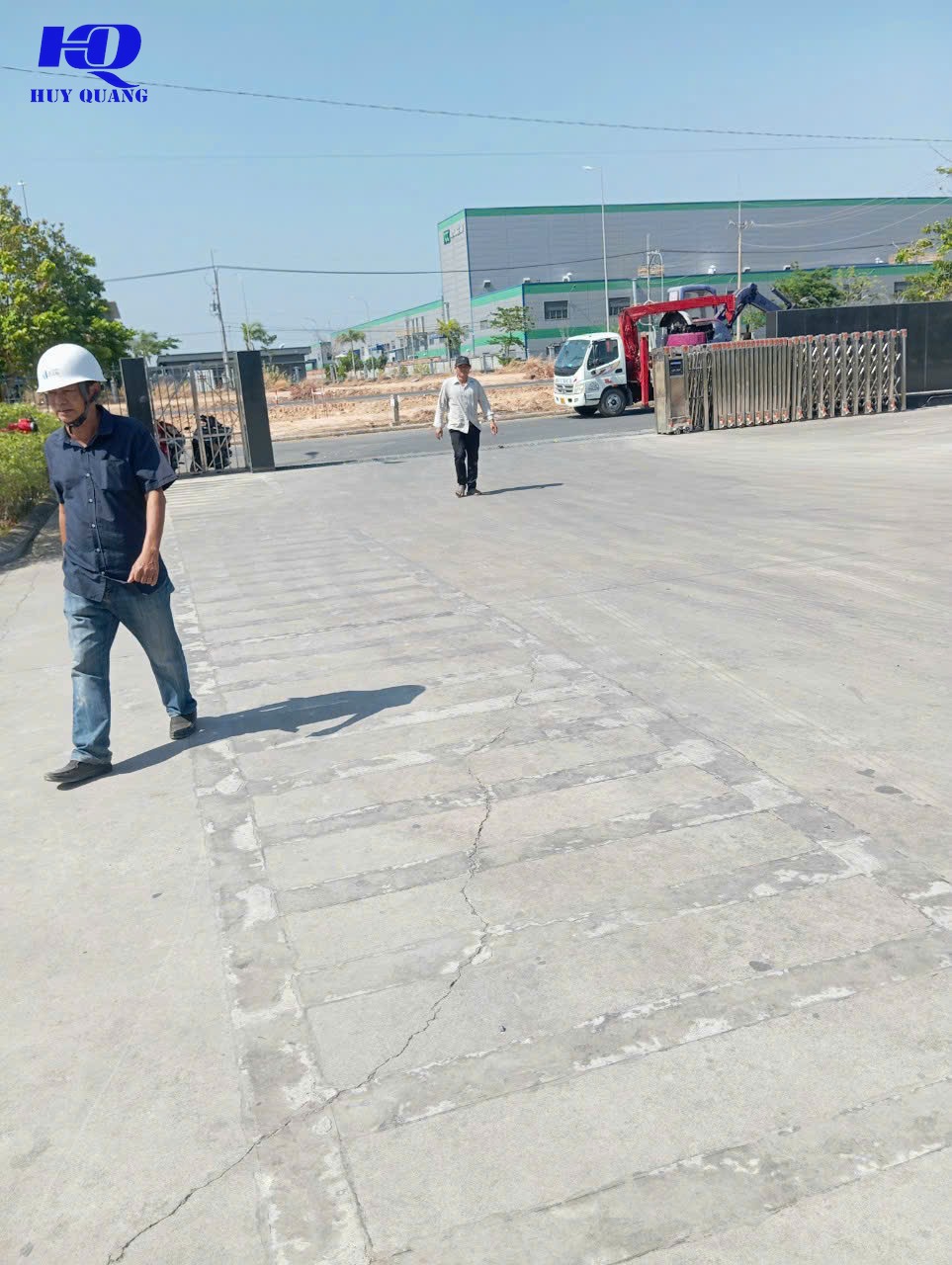 Hai nhân viên khảo sát đi trên nền bê tông nứt tại một mặt bằng khu công nghiệp, phía sau là cổng bảo vệ và xe cẩu nhỏ