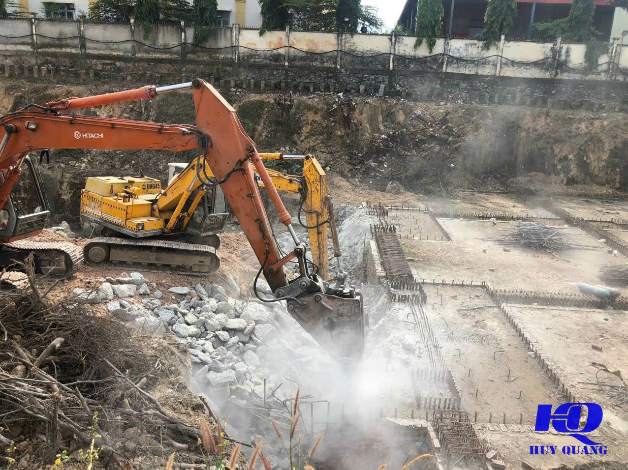 Máy đào gắn đầu đục thủy lực xử lý bê tông móng cũ, bụi bốc lên trong lúc thi công gần khung cốt thép mới