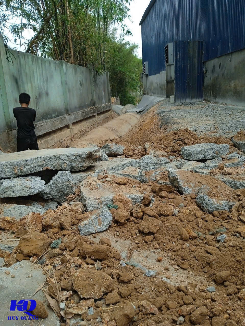 Rãnh đào lộ đường ống ngầm dọc tường bao nhà xưởng mái tôn xanh, các mảng bê tông cũ đã phá dỡ nằm rải rác cạnh rãnh
