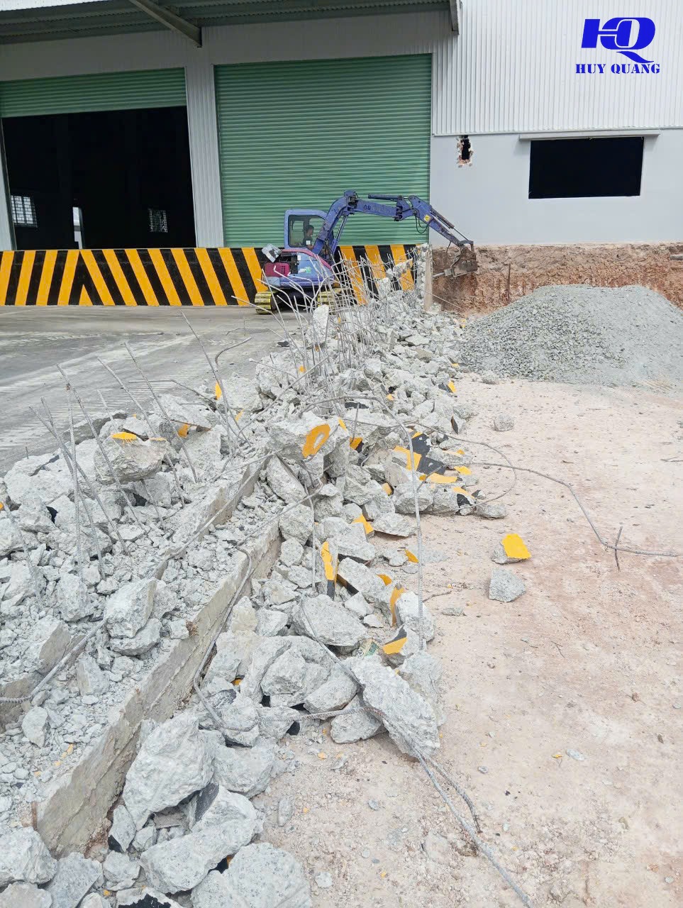 Xe múc mini gắn đầu đục thủy lực xử lý dải bê tông có cốt thép ngay trước cổng nhà xưởng có cửa cuốn màu xanh, đá base chờ sẵn để thi công lại