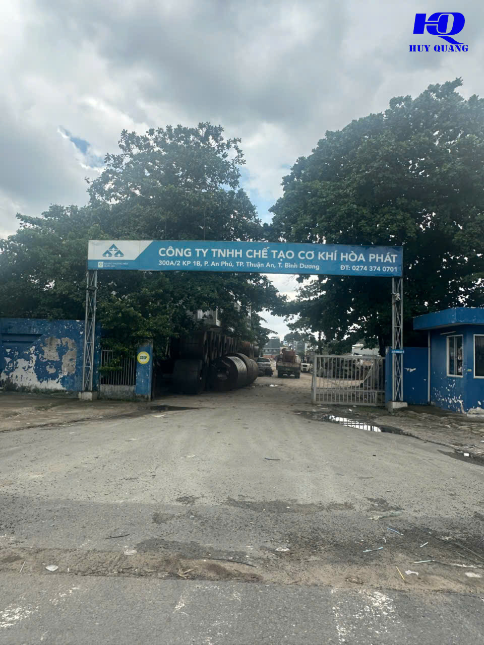 Cổng nhà máy với biển hiệu xanh, trong sân có cuộn thép và xe tải đậu cạnh khu vực sản xuất