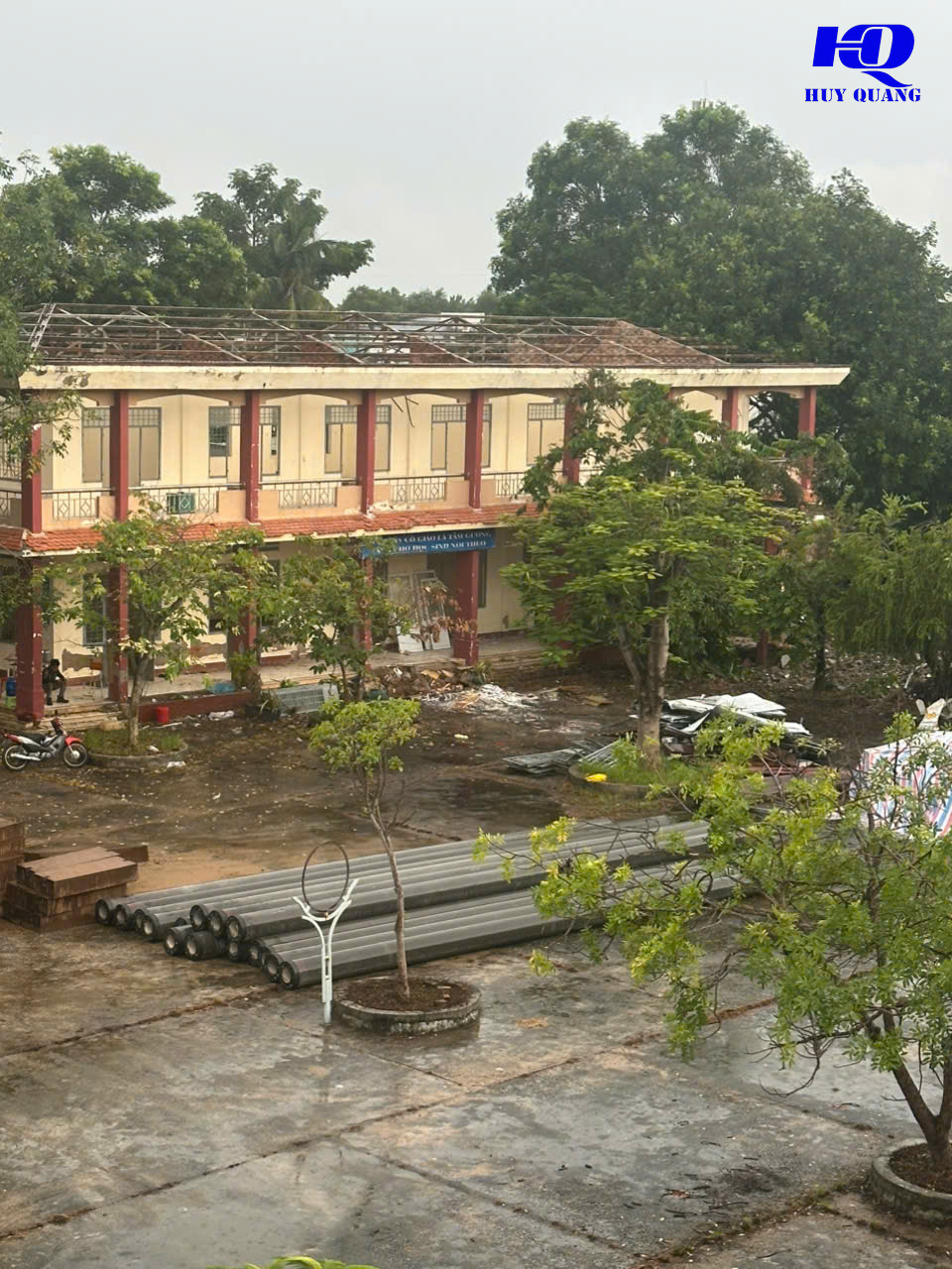 Dãy phòng học hai tầng nhìn từ trên cao, mái ngói đã dỡ hết chỉ còn khung kèo thép trần, sân trường tập kết ống cống bê tông